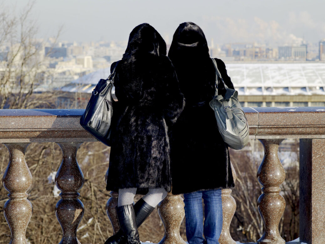 Two young women wearing black fur coats with handbags and hoods look out at a view in Moscow